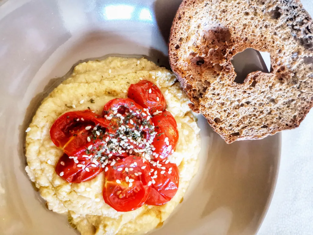 Bol de houmous garni de tomates rôties, graines de sésame et herbes, accompagné d’une tranche de pain grillé.