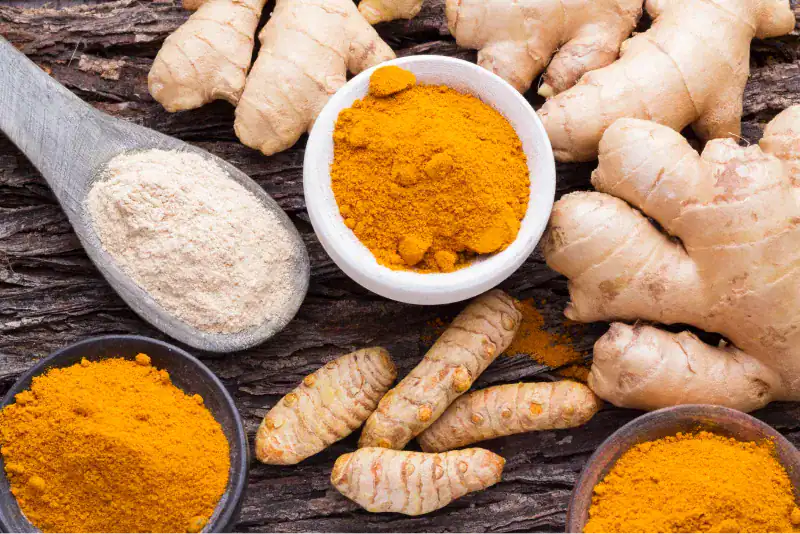 Racines et poudre de curcuma et de gingembre frais disposés sur une surface en bois rustique, avec des bols et une cuillère en bois remplis d'épices en poudre orange et beige