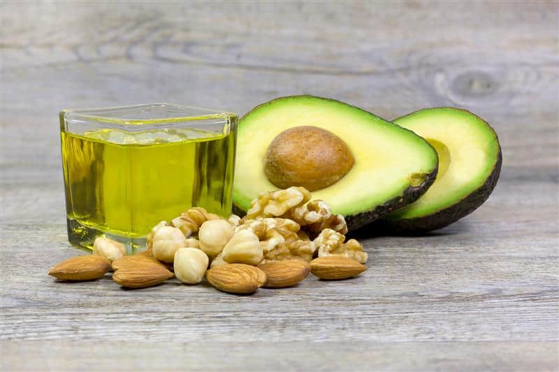 Aliments riches en graisses saines : avocat coupé en deux, mélange de noix, amandes et noisettes, et verre d'huile végétale dorée, disposés sur une surface en bois gris