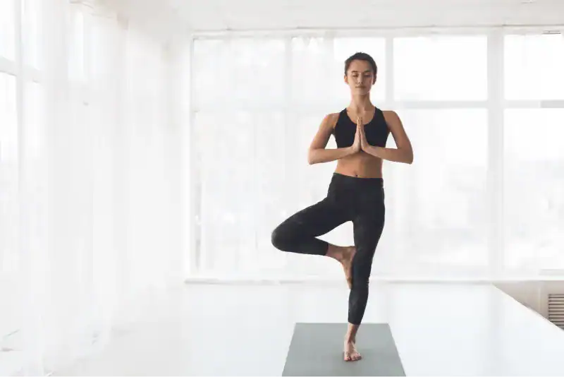 Femme en tenue de sport noire pratiquant la pose de l'arbre (Vrikshasana) sur un tapis de yoga gris, dans un studio lumineux aux grandes fenêtres