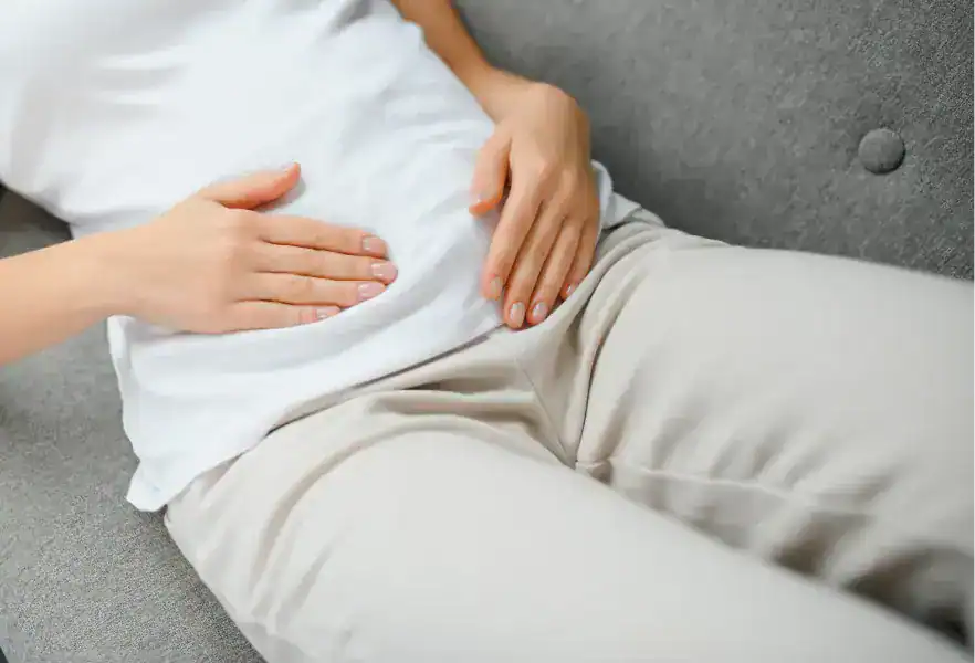 Personne allongée sur un canapé gris, les mains posées sur l'abdomen, en t-shirt blanc et pantalon beige
