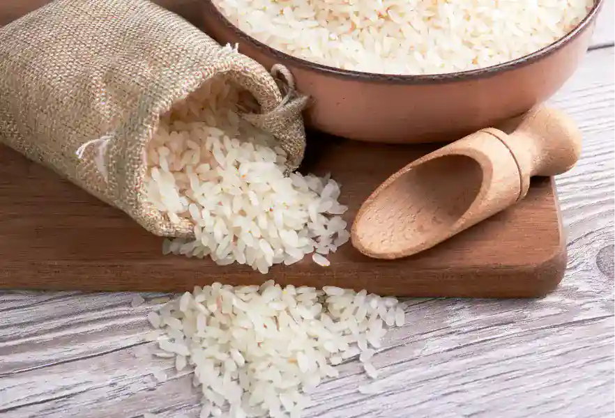 Riz blanc cru versé d'un sac en jute sur une planche en bois, avec un bol en cuivre et une cuillère en bois, sur surface en bois clair