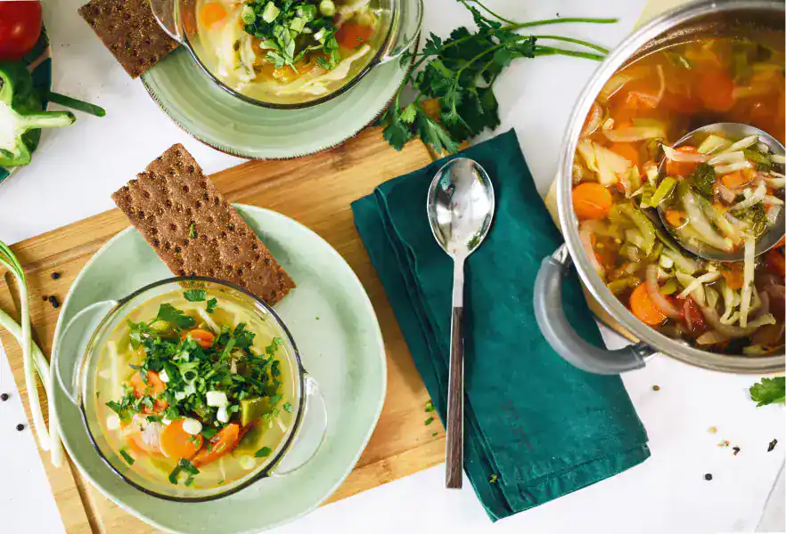 Soupe de légumes maison aux carottes, poireaux et persil frais, servie dans des bols en verre sur une planche en bois, avec une casserole et des crackers de seigle