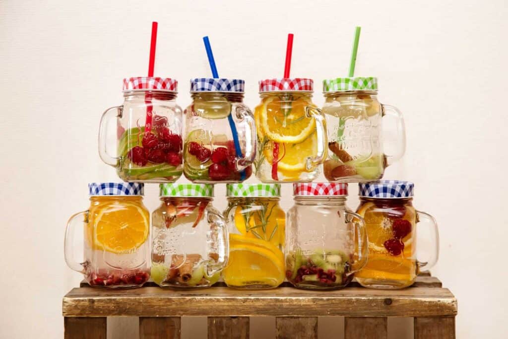 Neuf bocaux en verre avec couvercles à carreaux colorés et pailles, remplis d'eaux aromatisées aux fruits : framboises, orange, kiwi, romarin et grenade, disposés sur une palette en bois